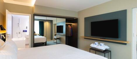 A hotel room with a large bed, a TV mounted on the wall, a bathroom with a shower, and a mirror.