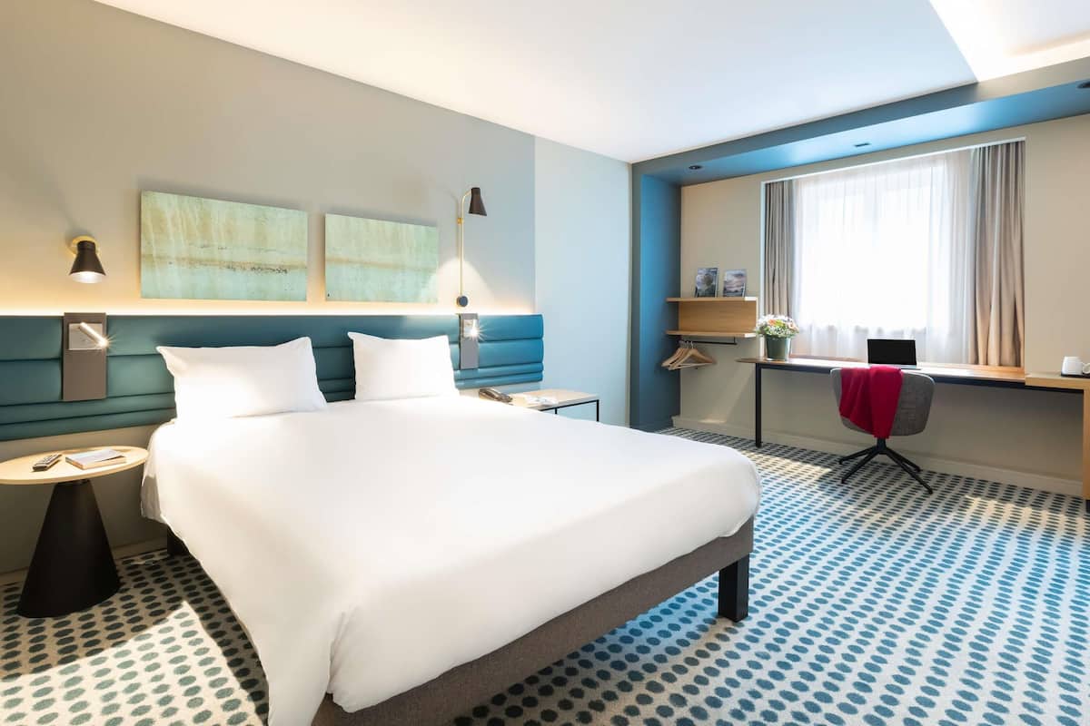 A hotel room with a large bed, a desk with a chair, a window with curtains, and artwork on the wall.