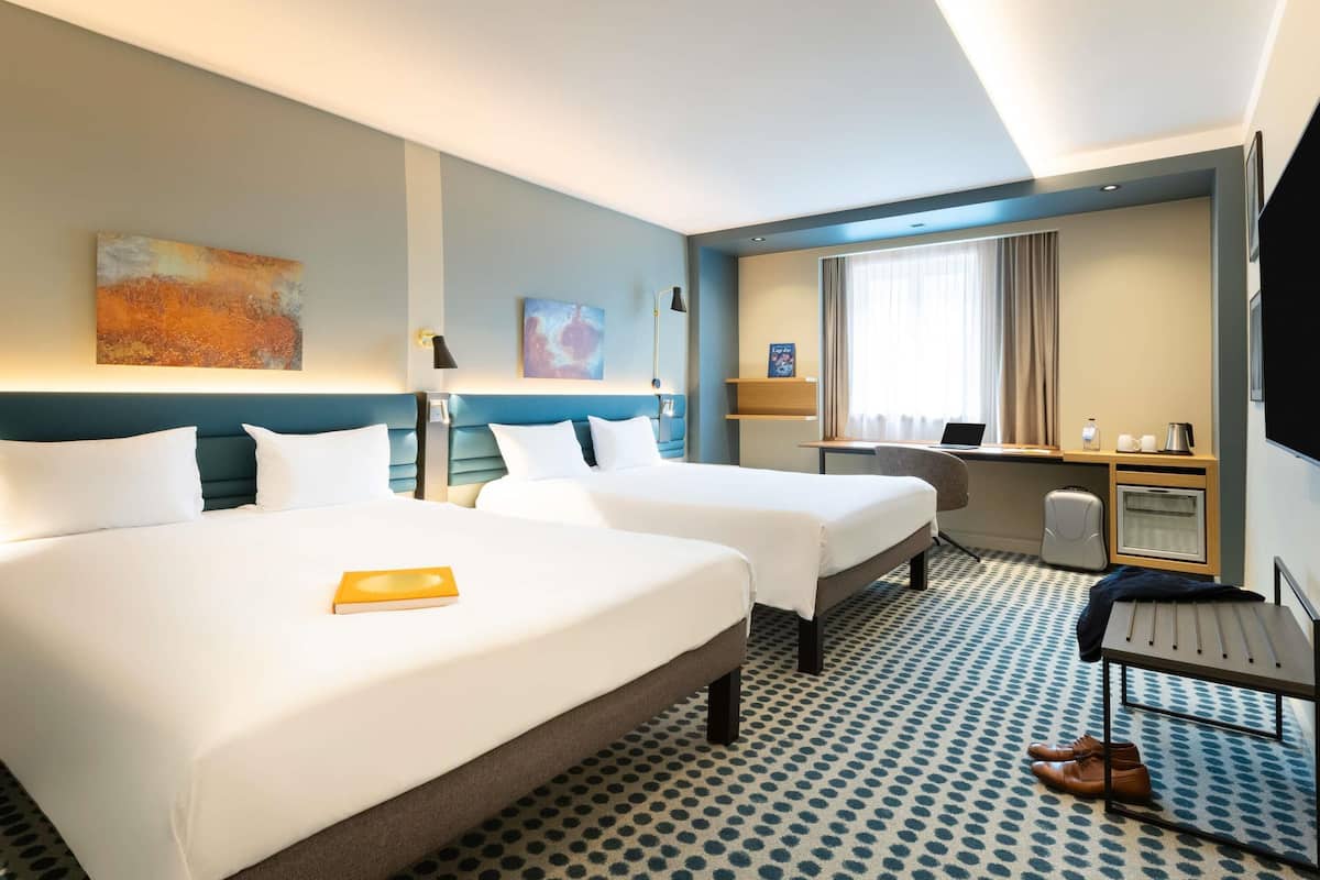 A hotel room with two beds, a desk, a chair, a TV, and a small kitchenette.