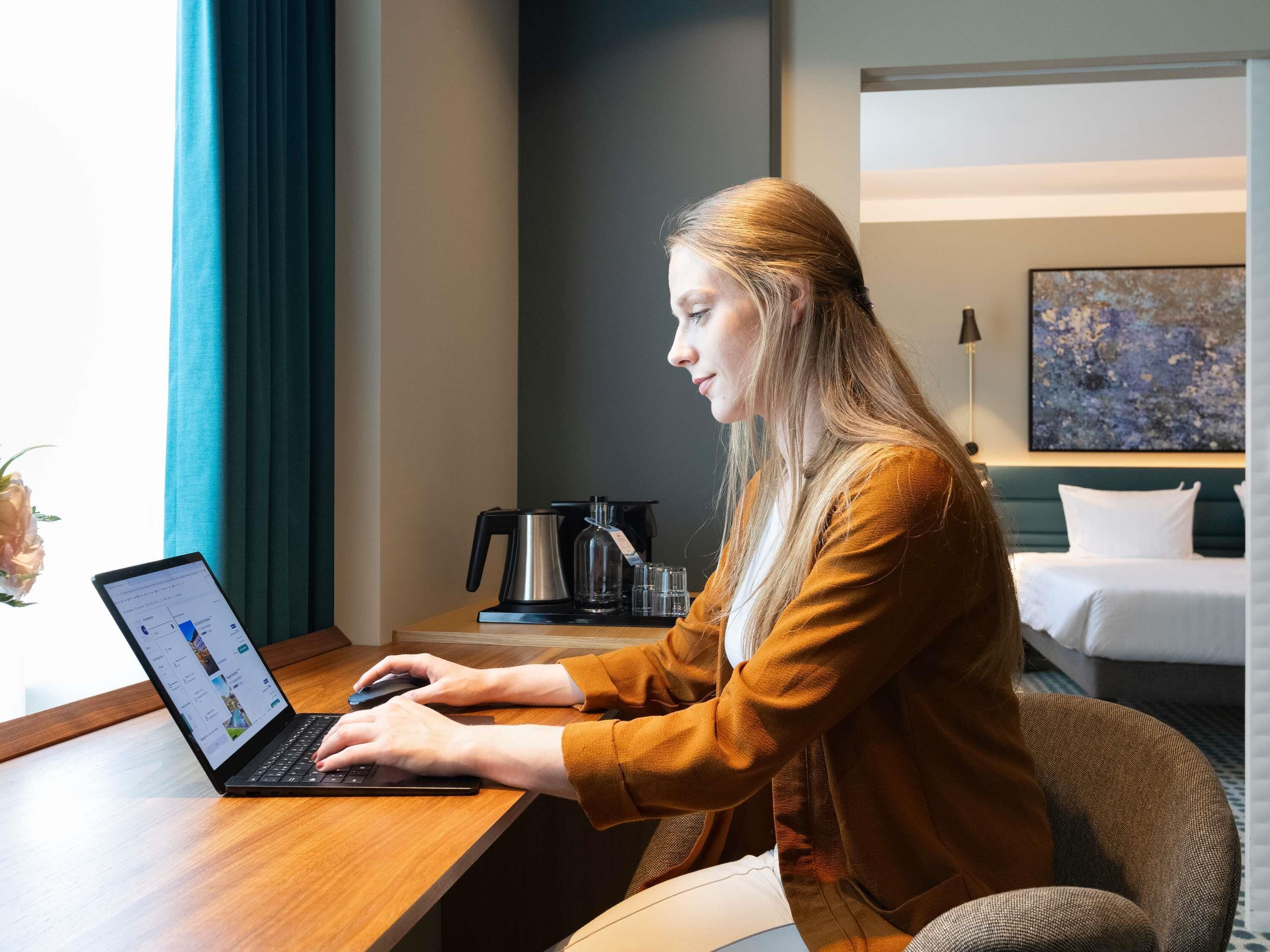 Uma pessoa trabalhando em um laptop em quarto de hotel, com uma cama, uma cafeteira e um quadro na parede.