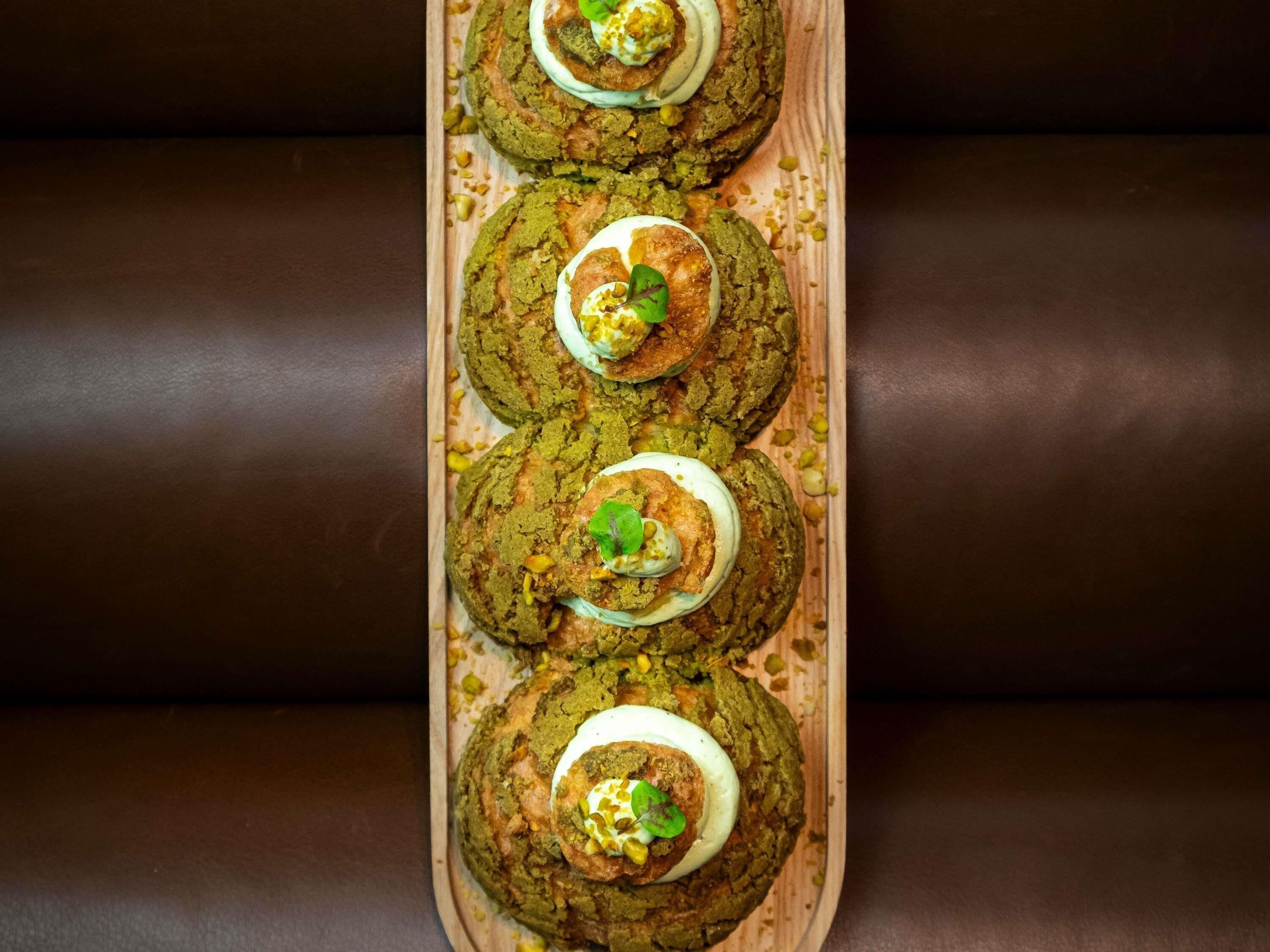 Una bandeja de madera con cuatro galletas verdes cubiertas de crema blanca y una hoja verde, sobre una superficie de cuero marrón oscuro.