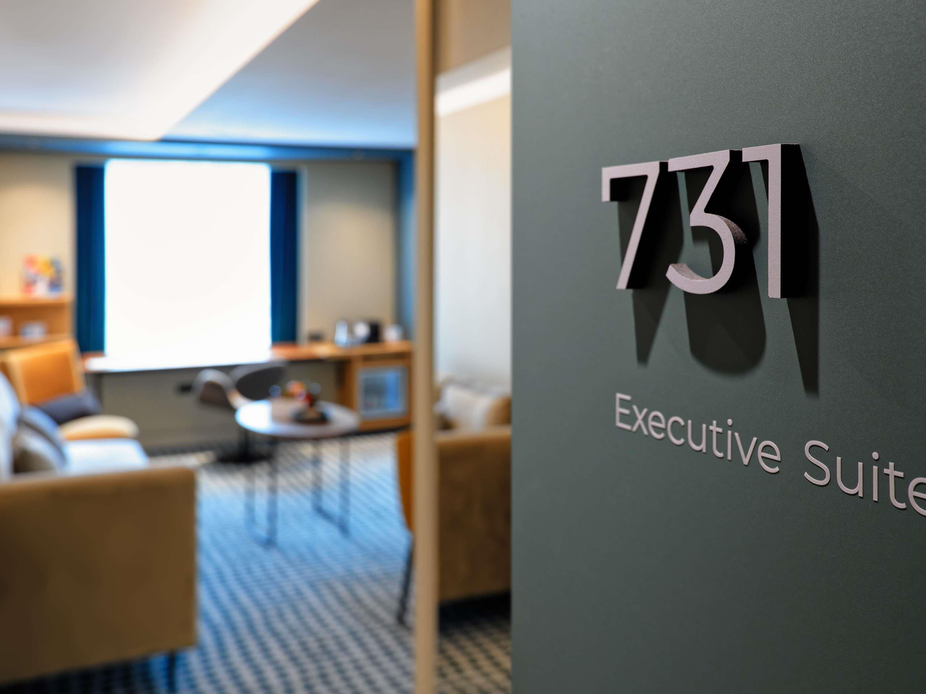 Une chambre d’hôtel moderne dotée d’une grande fenêtre, d’un canapé, d’une petite table et d’une pancarte indiquant « Suite Exécutive 731 ».