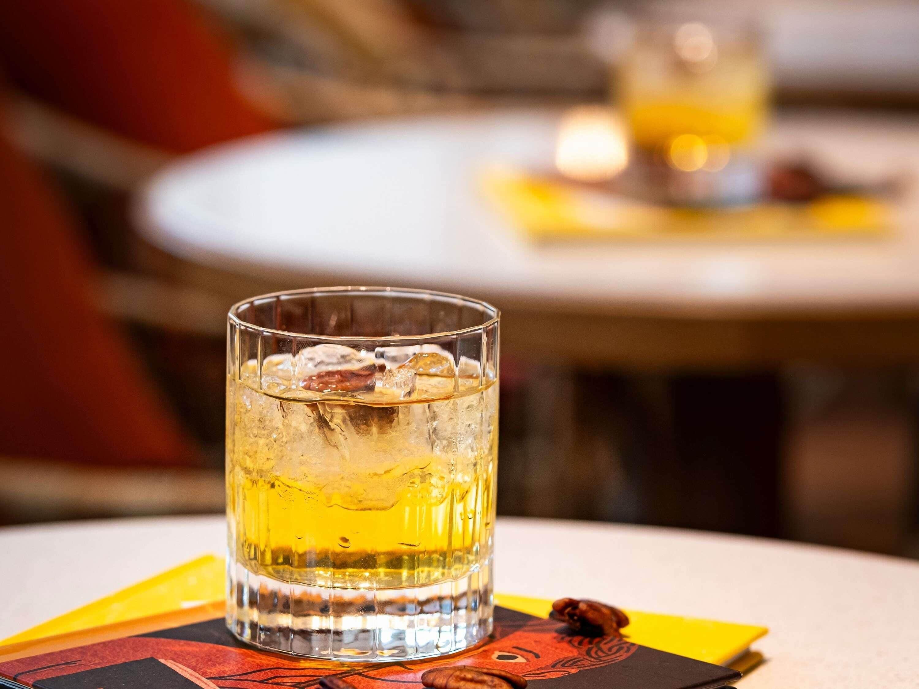 A glass of amber-colored beverage with ice cubes on a coaster, placed on a table with a blurred background.