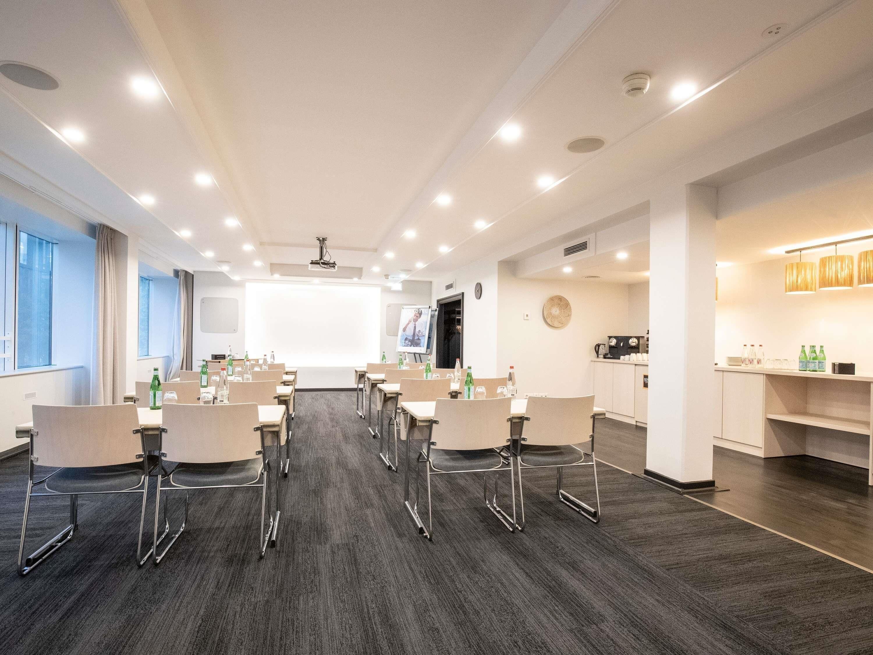 Un salón de conferencias con una pantalla grande, mesas, sillas y una zona de kitchenette.