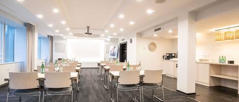 Un salón de conferencias con una pantalla grande, mesas, sillas y una zona de kitchenette.