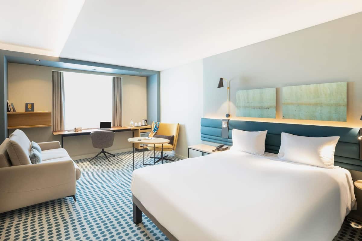 A modern hotel room with a large bed, a desk, a chair, and a sofa.