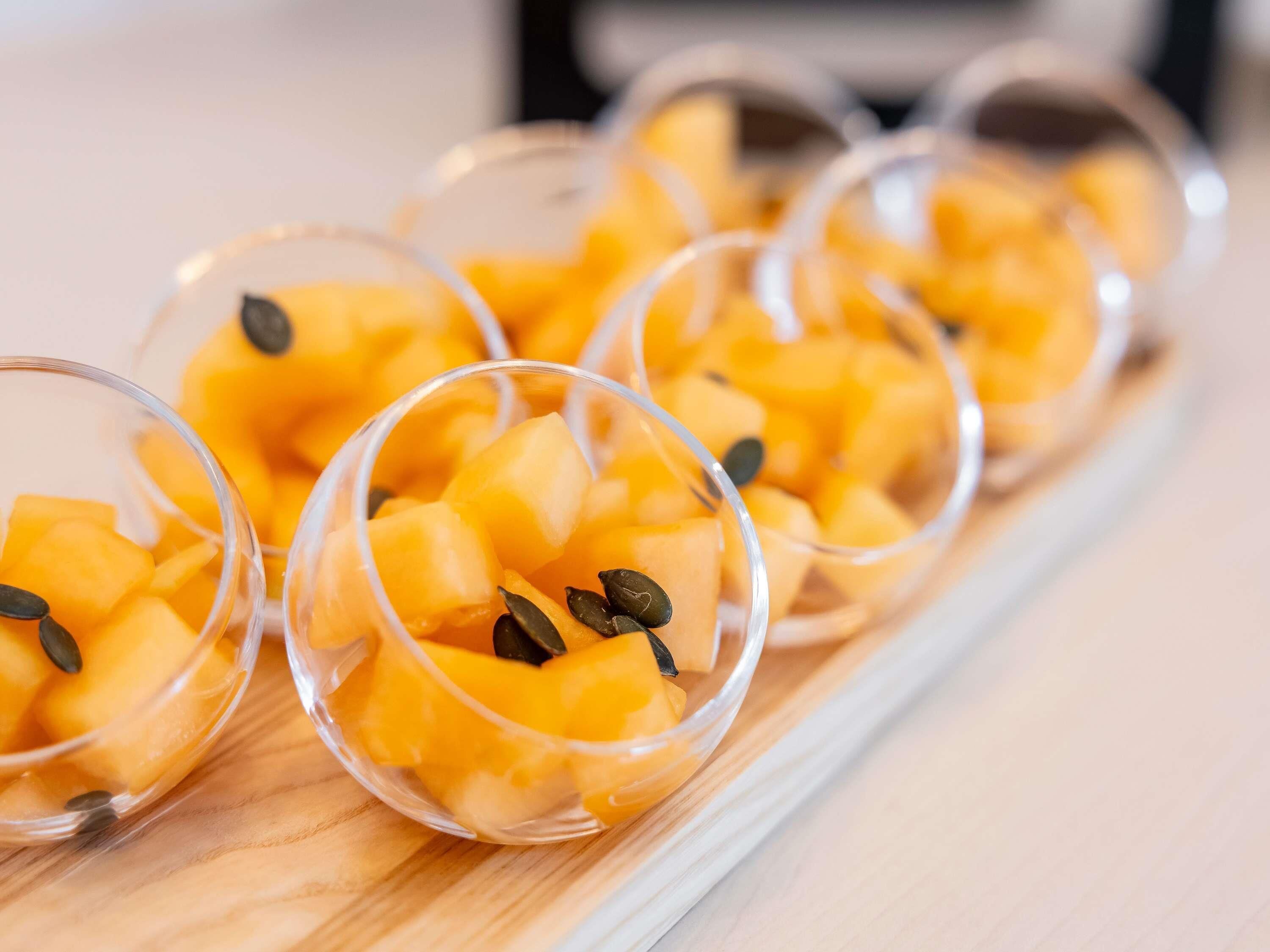 A series of clear glass bowls containing diced mango and pumpkin seeds arranged on a wooden surface.