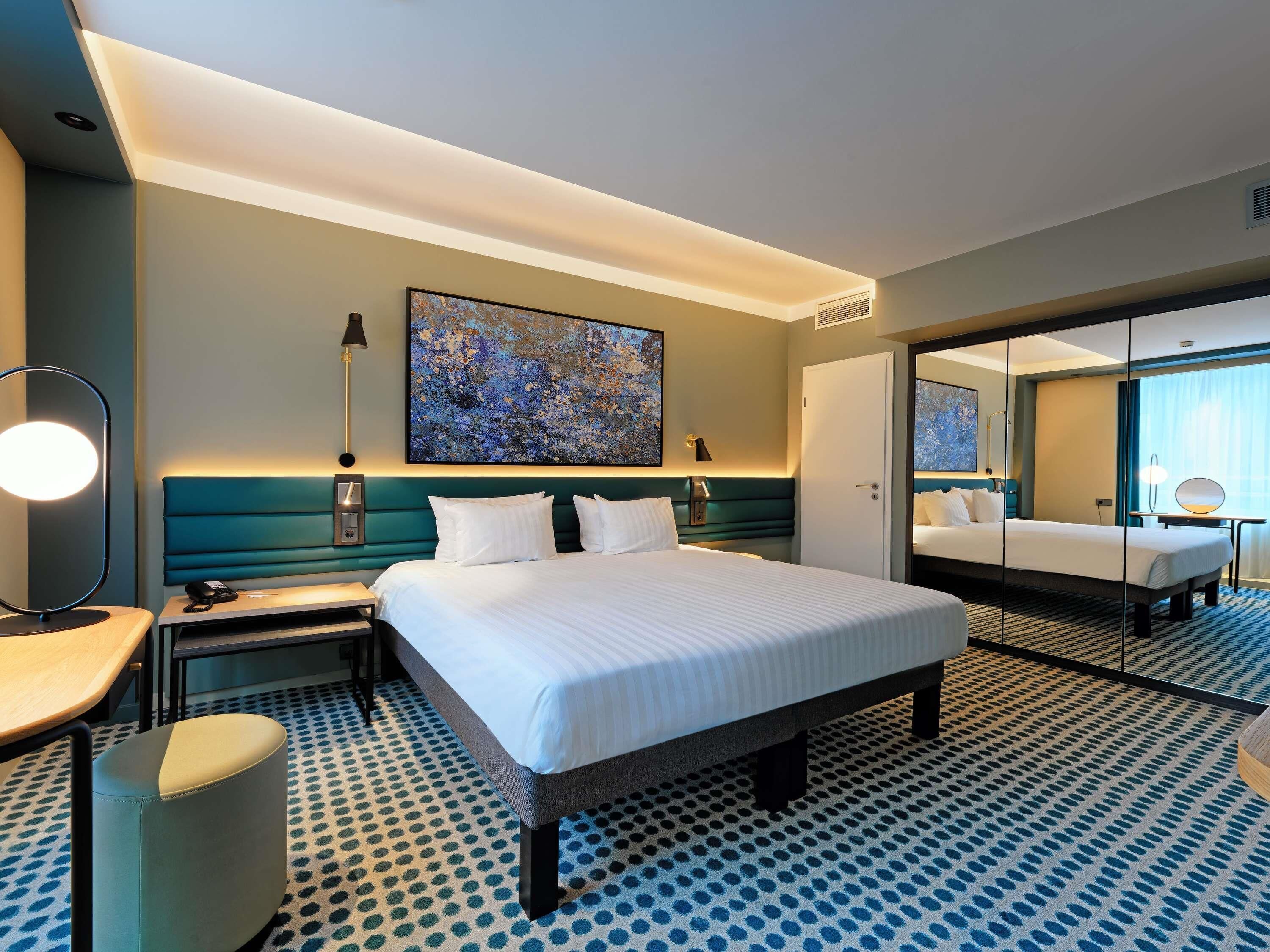 A hotel room with a large bed, a desk, a chair, a lamp, a painting, and a mirror.