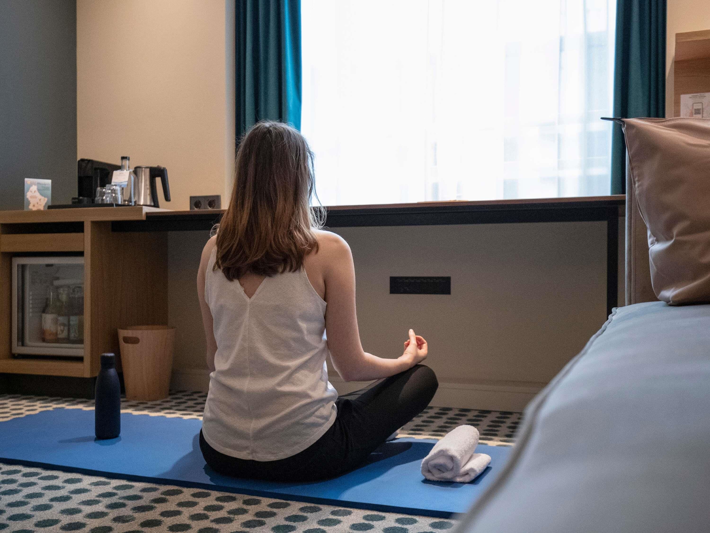 A person sitting on a blue exercise mat in a room with a bed, a small table with a kettle, and a window with curtains.