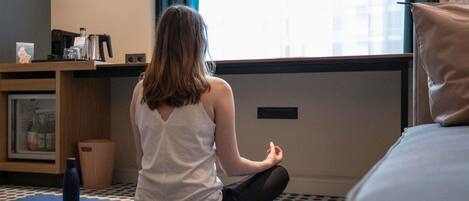 A person sitting on a blue exercise mat in a room with a bed, a small table with a kettle, and a window with curtains.