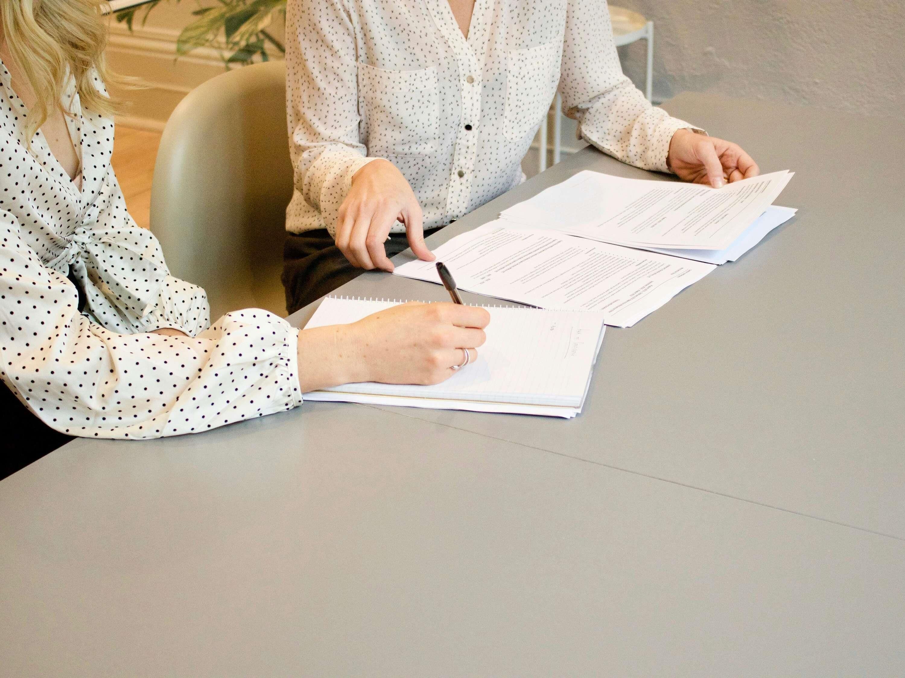 Two individuals seated at a table with documents, one signing a document.