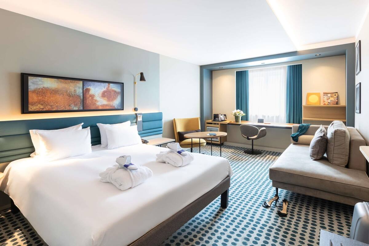 A hotel room with a bed, a desk, a chair, a sofa, a window with curtains, and artwork on the wall.