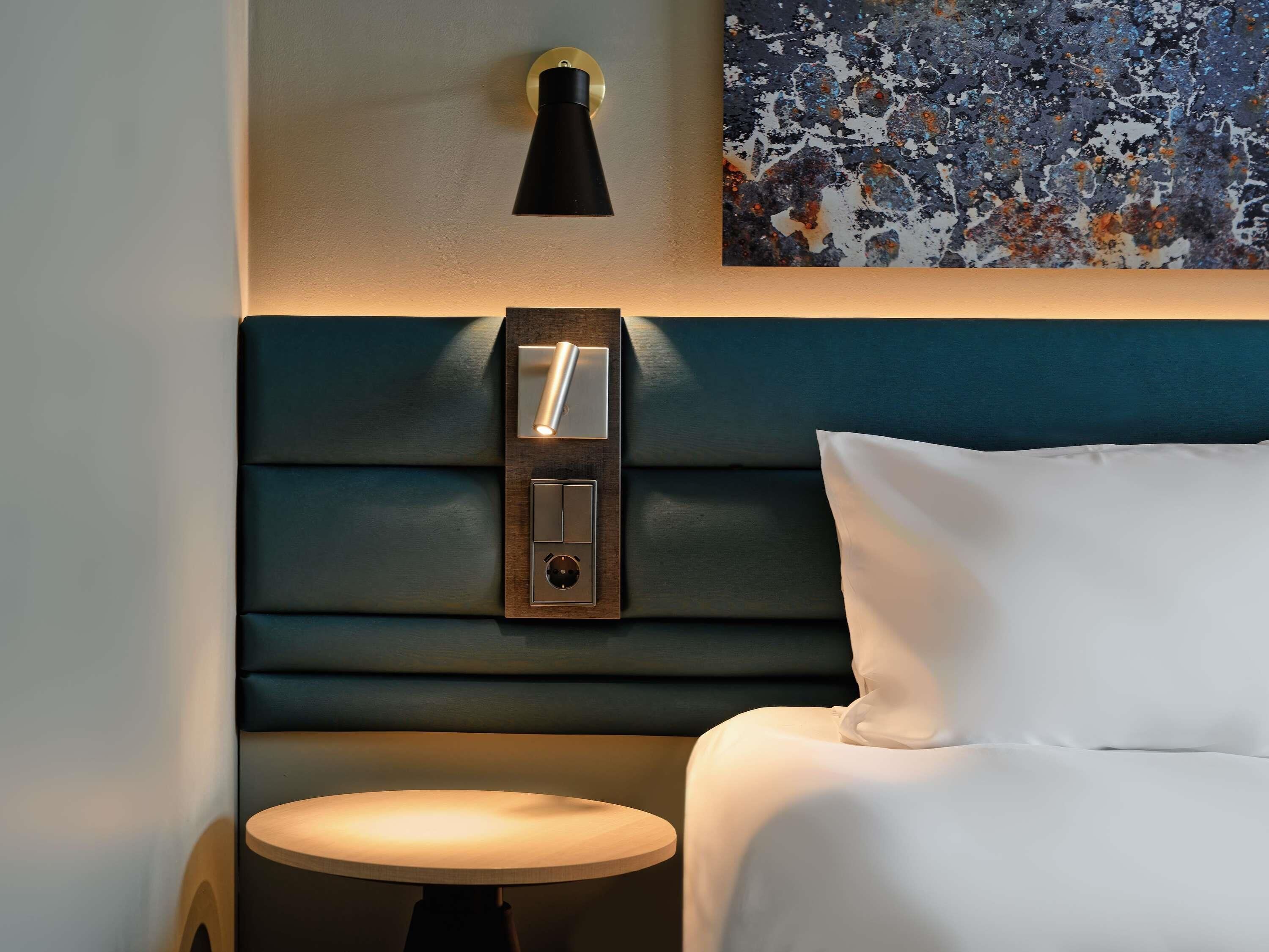 Een hotelkamer met een bed, nachtkastje, wandlamp en een schilderij.