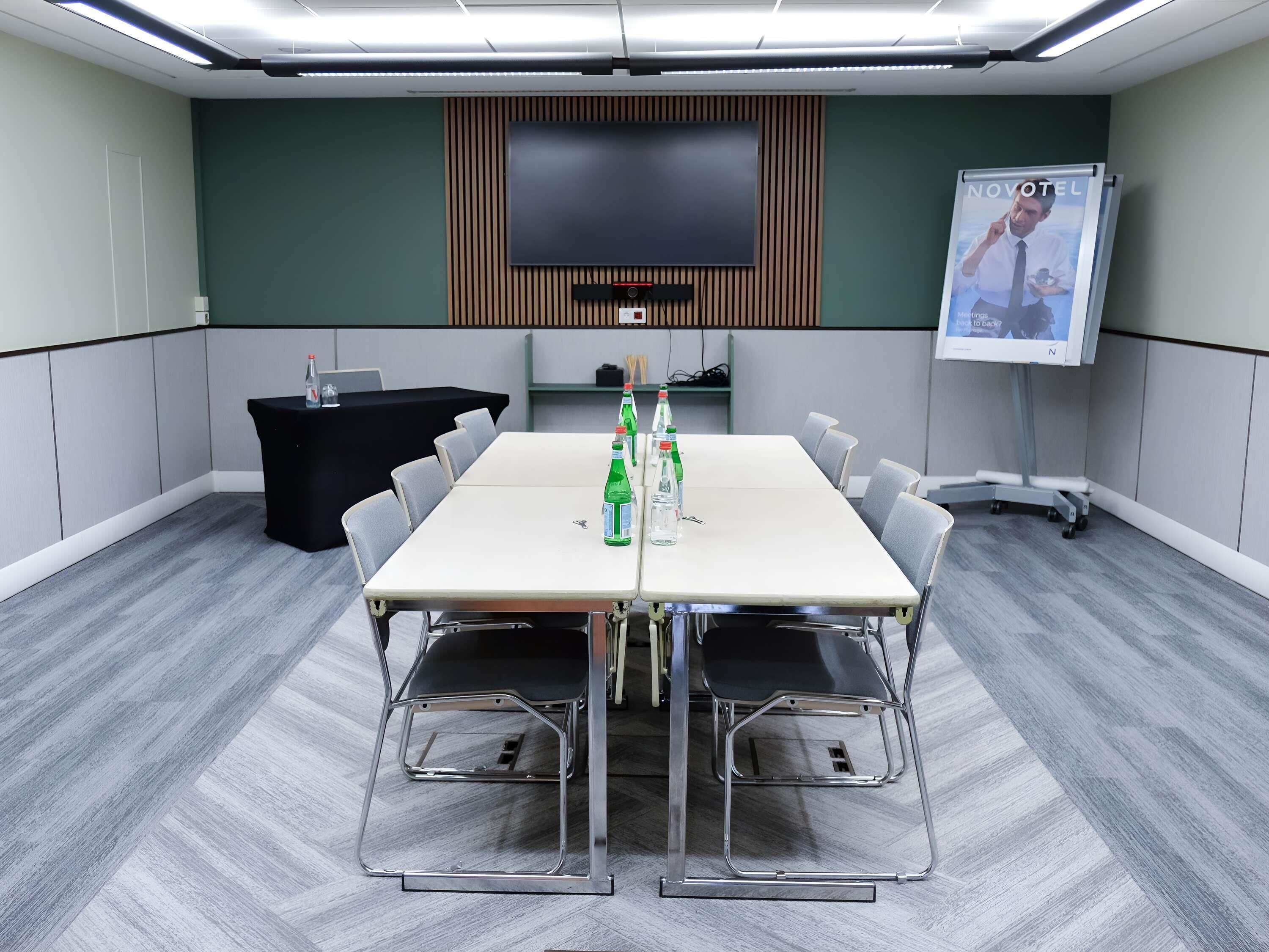 Un salón de conferencias con una larga mesa, sillas, un atril, una pantalla y un cartel.