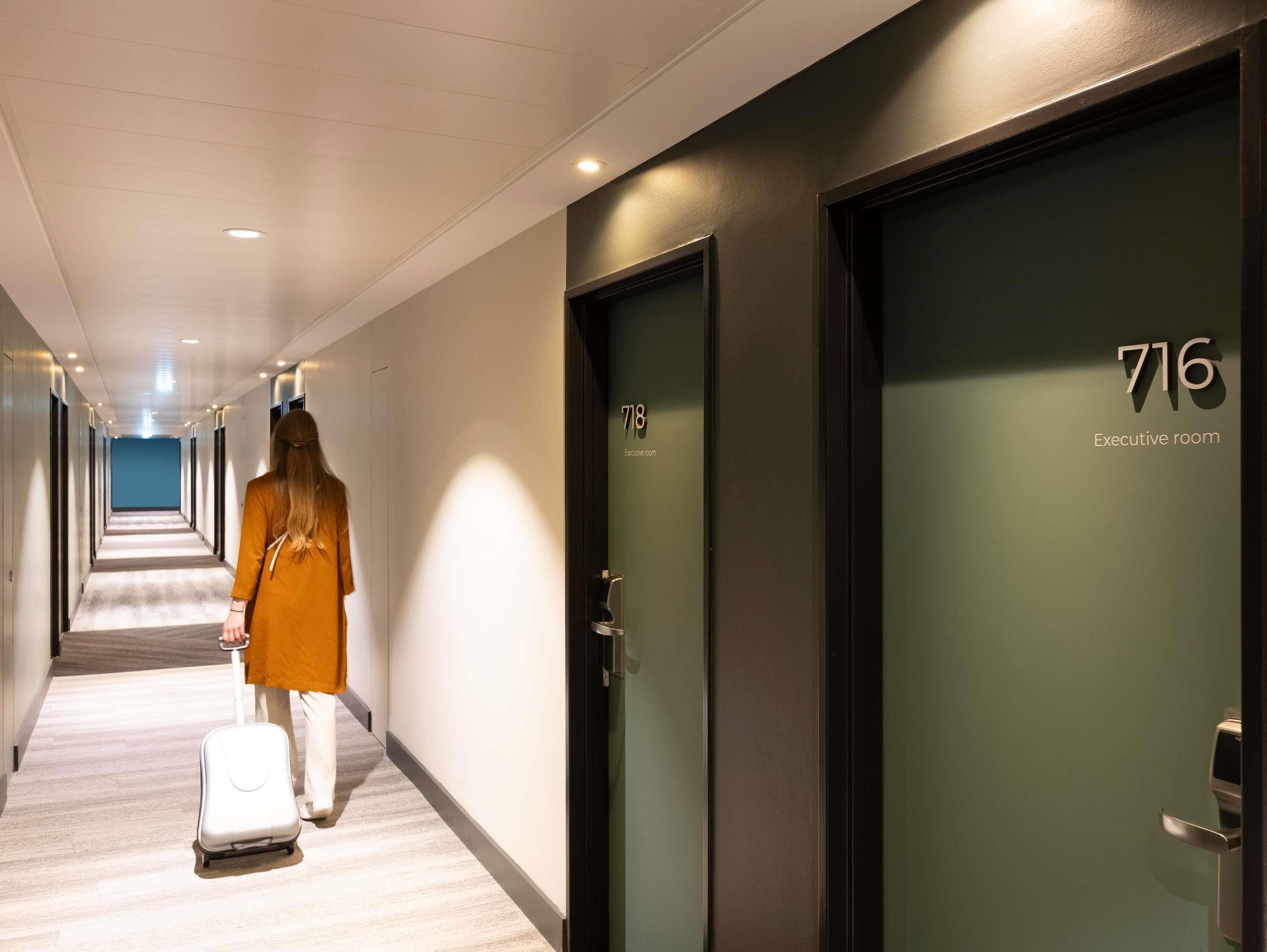 A hotel corridor with a person walking with a suitcase, doors numbered 718 and 716, and an "Executive room" sign.