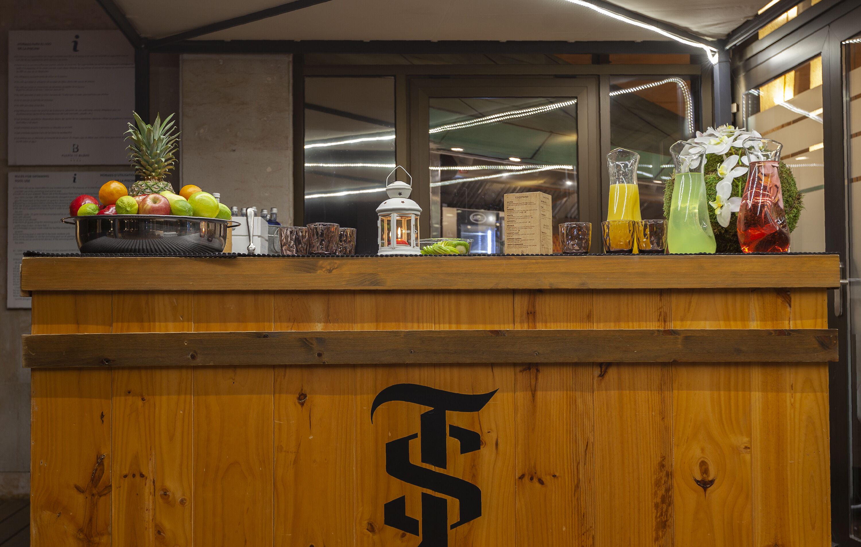 A wooden counter with a logo, a fruit bowl, a lantern, and three glass bottles with different colored liquids.