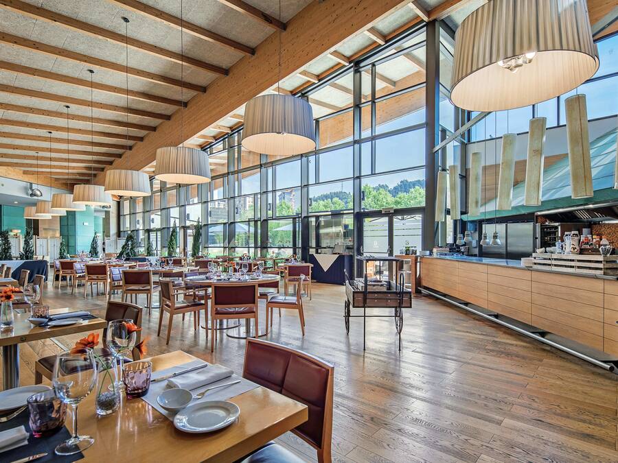 Un restaurante espacioso con pisos de madera y ventanas amplias, que ofrece vistas al exterior. Las mesas están dispuestas con platos y cubiertos, y hay lámparas colgantes.