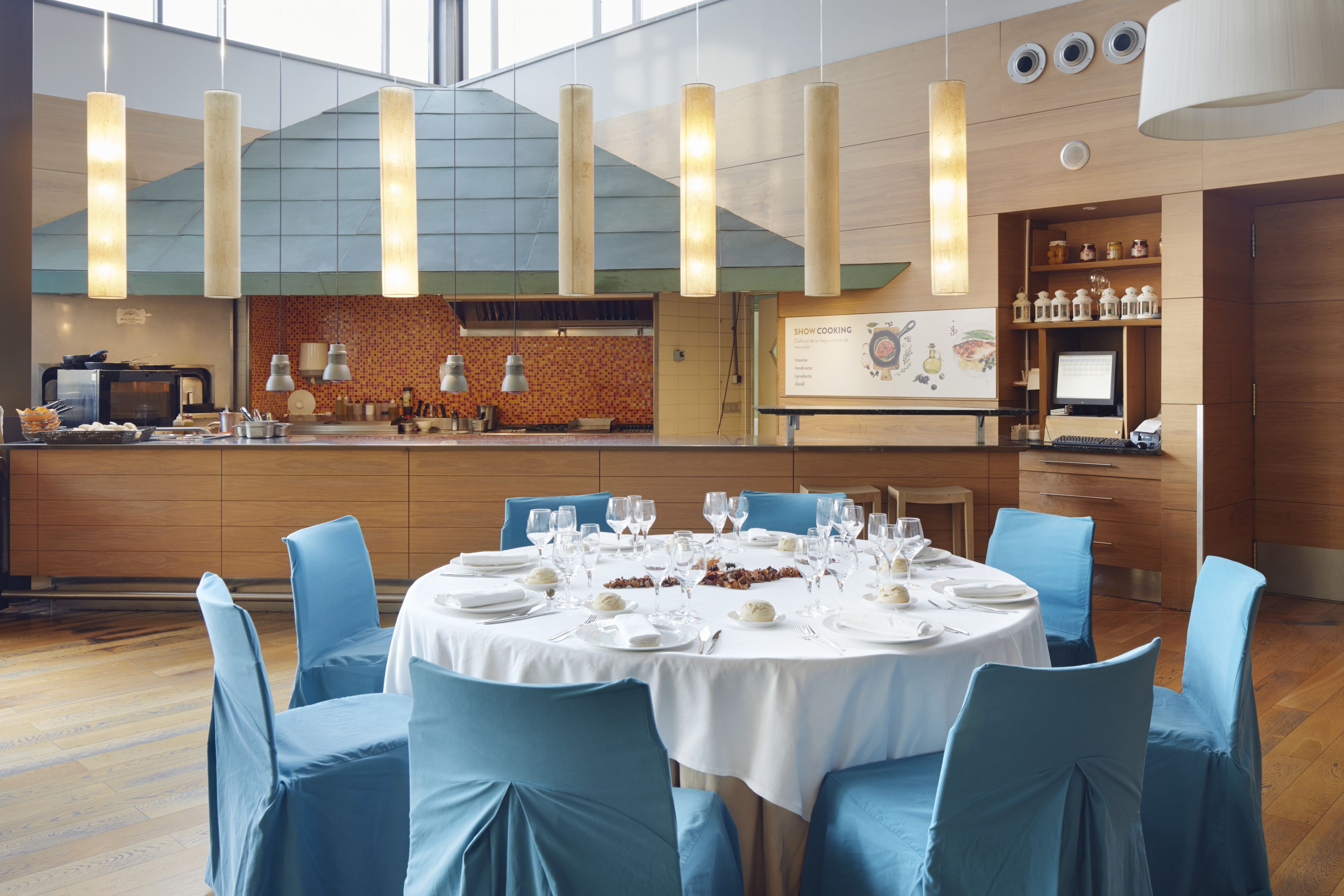 A modern dining area with a round table set for six, blue chairs, and a kitchen visible in the background.
