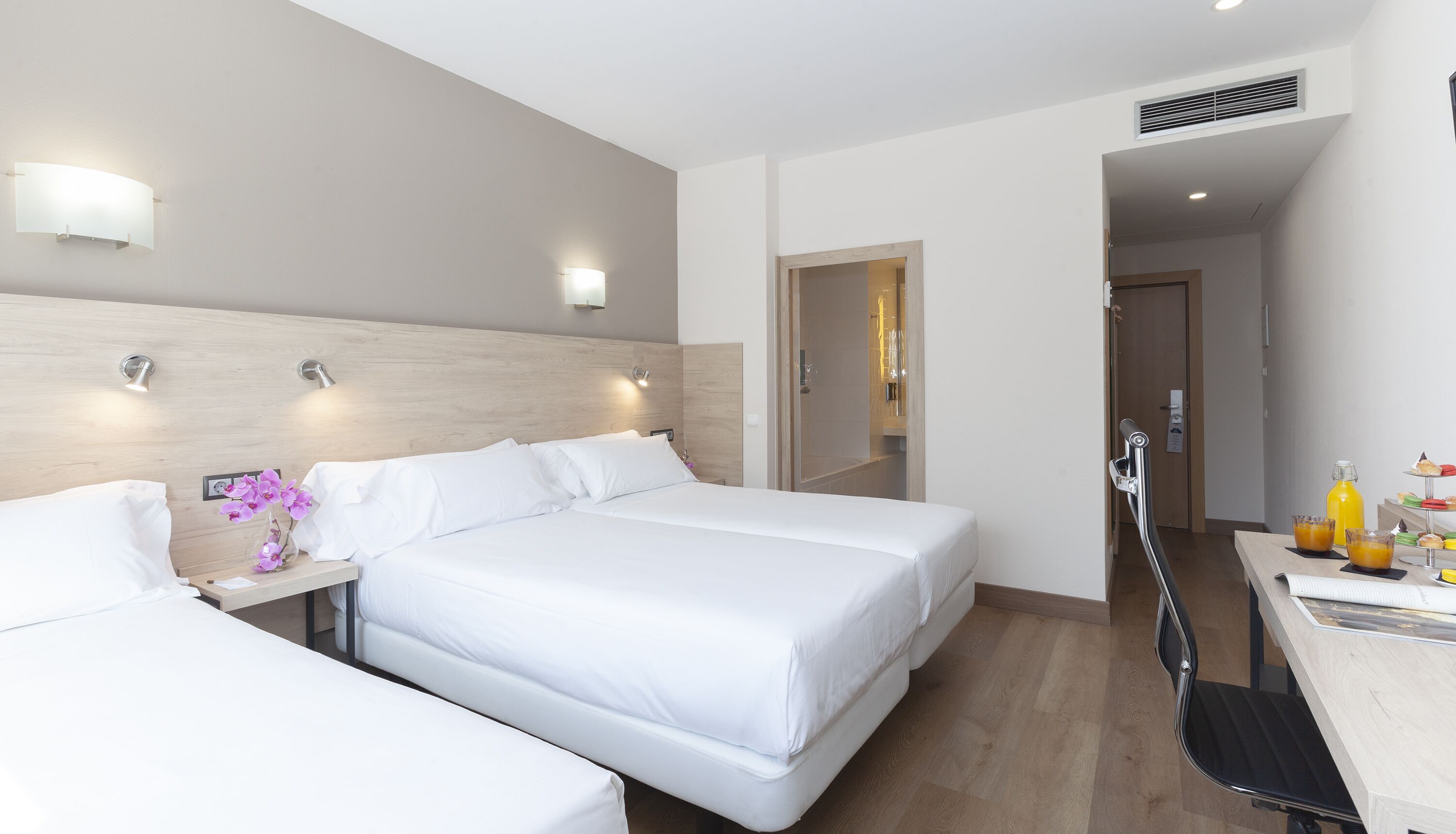 Une chambre d’hôtel avec deux lits simples, un bureau avec une chaise, une petite table avec une bouteille et des verres, et une salle de bain visible à travers une porte ouverte.