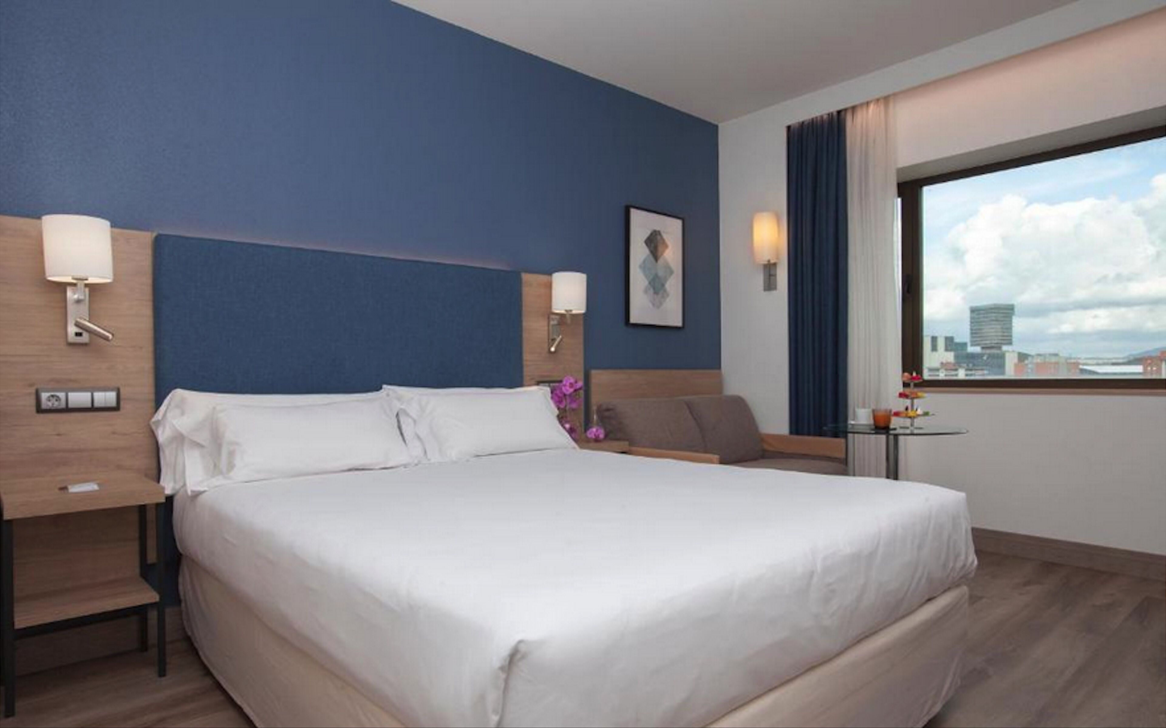 A hotel room with a large bed, two bedside tables, a small table with a drink, and a window with a city view.
