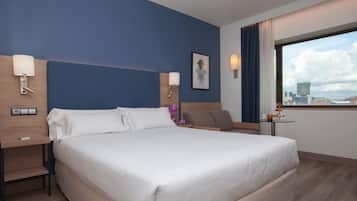 A hotel room with a large bed, two bedside tables, a small table with a drink, and a window with a city view.
