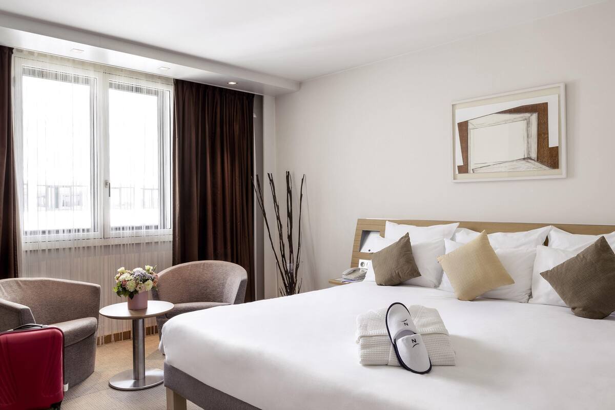 A hotel room with a large bed, a desk, two chairs, a window with curtains, and a framed picture on the wall.