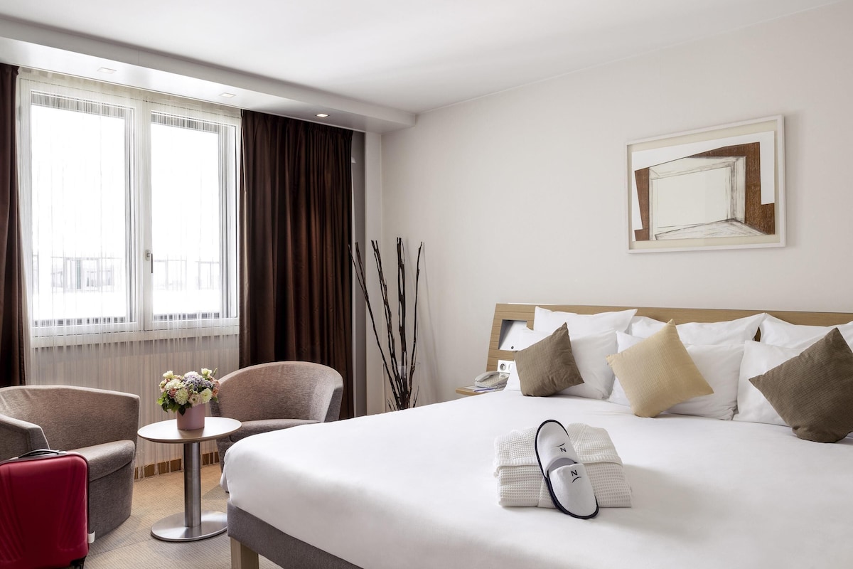 A hotel room with a large bed, a desk, two chairs, a window with curtains, and a framed picture on the wall.