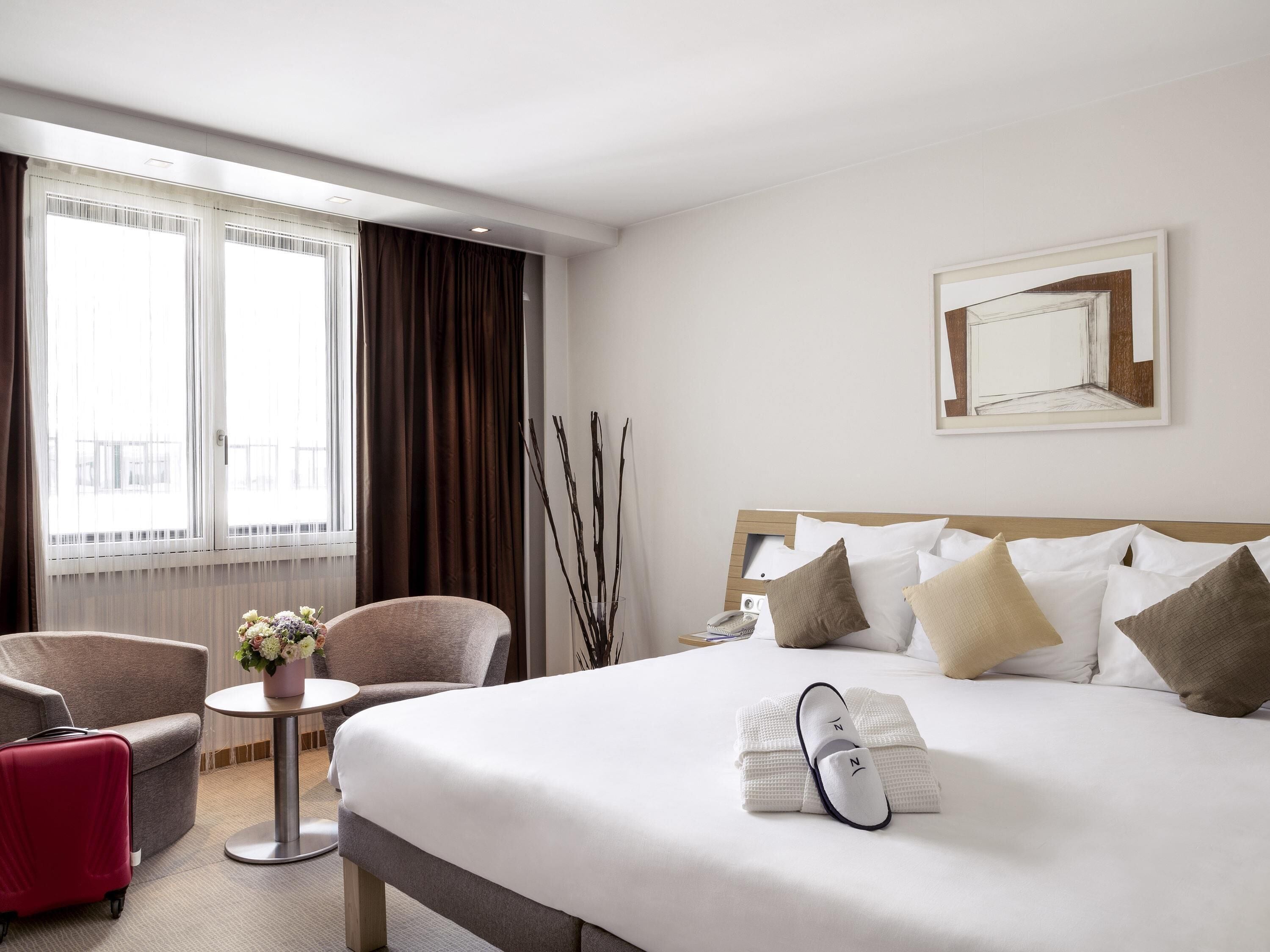 A hotel room with a large bed, a desk, two chairs, a window with curtains, and a framed picture on the wall.