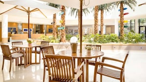 A spacious indoor dining area with wooden tables and chairs, large umbrellas, and a central planter with greenery. - Novotel Paris Roissy CDG Convention (Roissy-en-France)