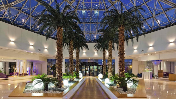 A spacious lobby with a high ceiling and a glass dome, featuring palm trees and a central water feature. - Novotel Paris Roissy CDG Convention (Roissy-en-France)