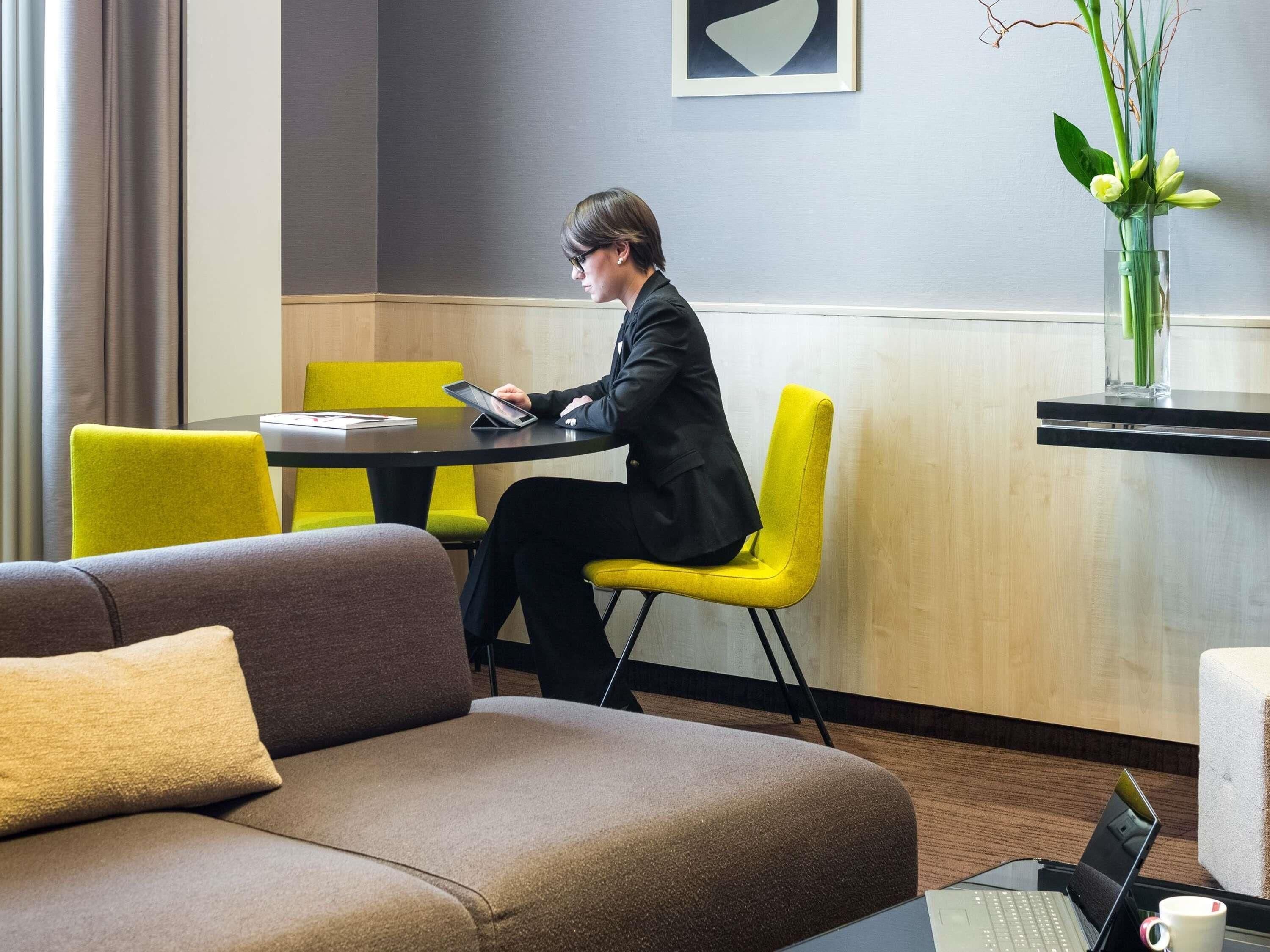 Une chambre d’hôtel moderne avec un canapé, une table à manger avec des chaises, un ordinateur portable posé sur un support et un vase rempli de fleurs.