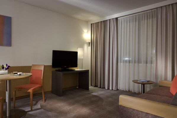 Une chambre d’hôtel avec une télévision, une petite table, un canapé et une fenêtre avec des rideaux.