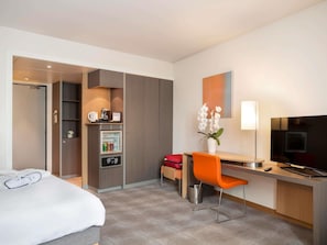A hotel room with a bed, a desk with a chair, a television, a minibar, and a small refrigerator. - Novotel Paris Roissy CDG Convention (Roissy-en-France)