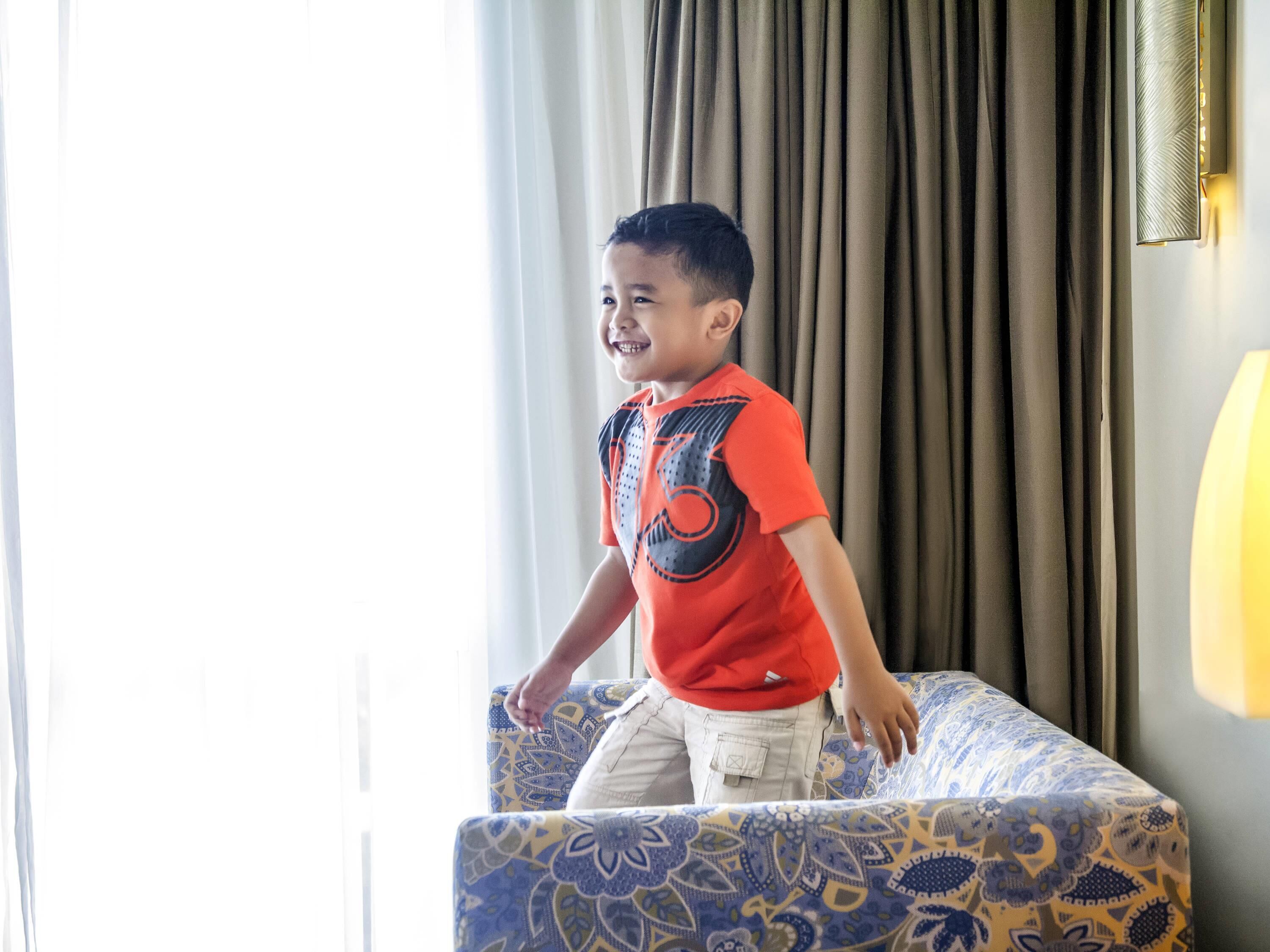 A child standing on a patterned sofa in a room with curtains and a wall-mounted light fixture.