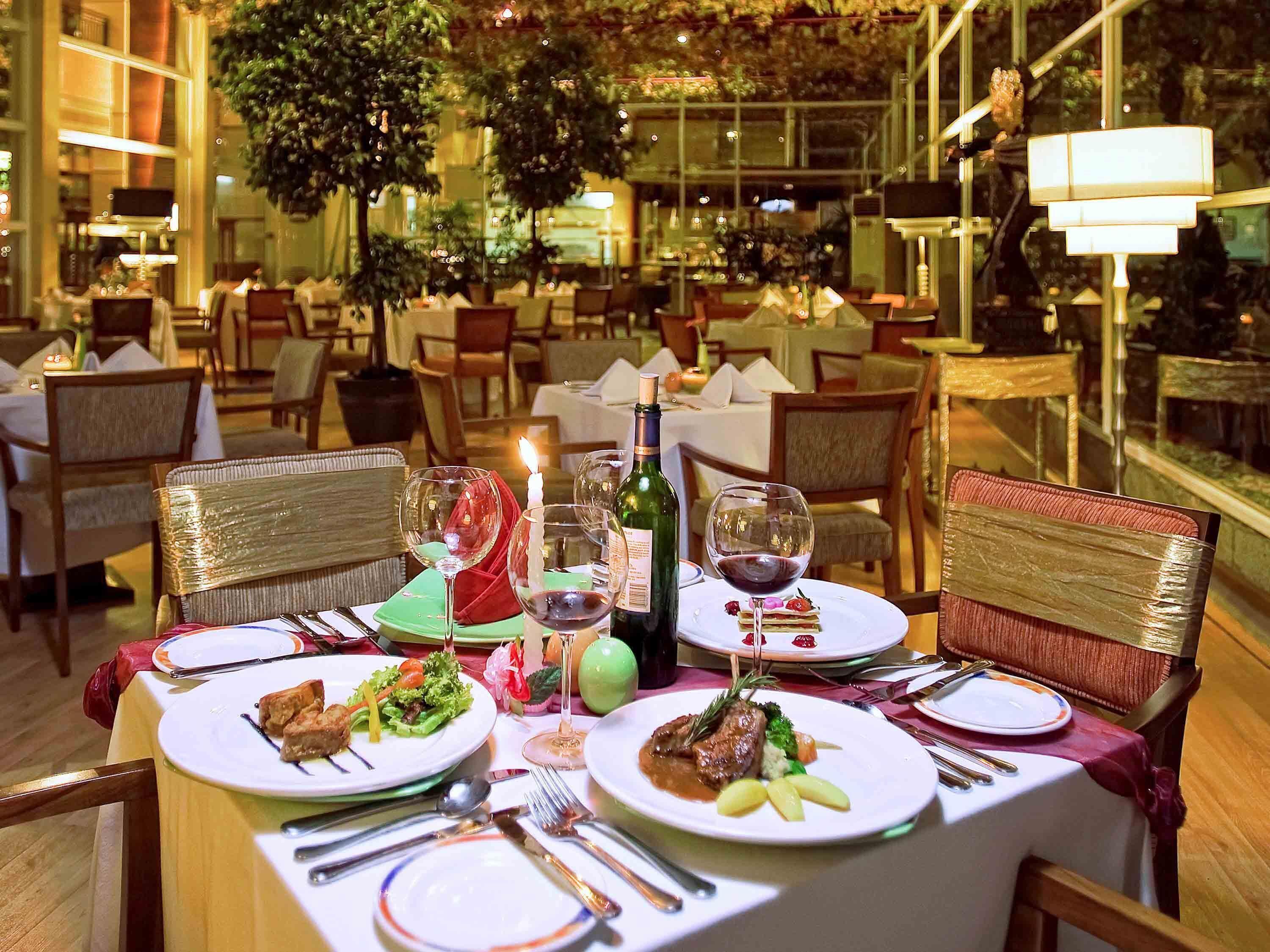 Une scène de restaurant, avec des tables dressées pour le service, où l’on voit des assiettes de nourriture, des verres à vin et des couverts.