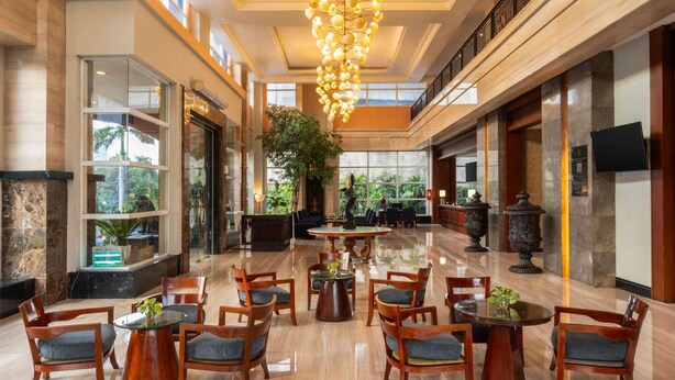 A spacious lobby with a high ceiling, large windows, and a central chandelier. There are multiple seating areas with tables and chairs, and a reception desk is visible in the background. - Novotel Semarang (Semarang)