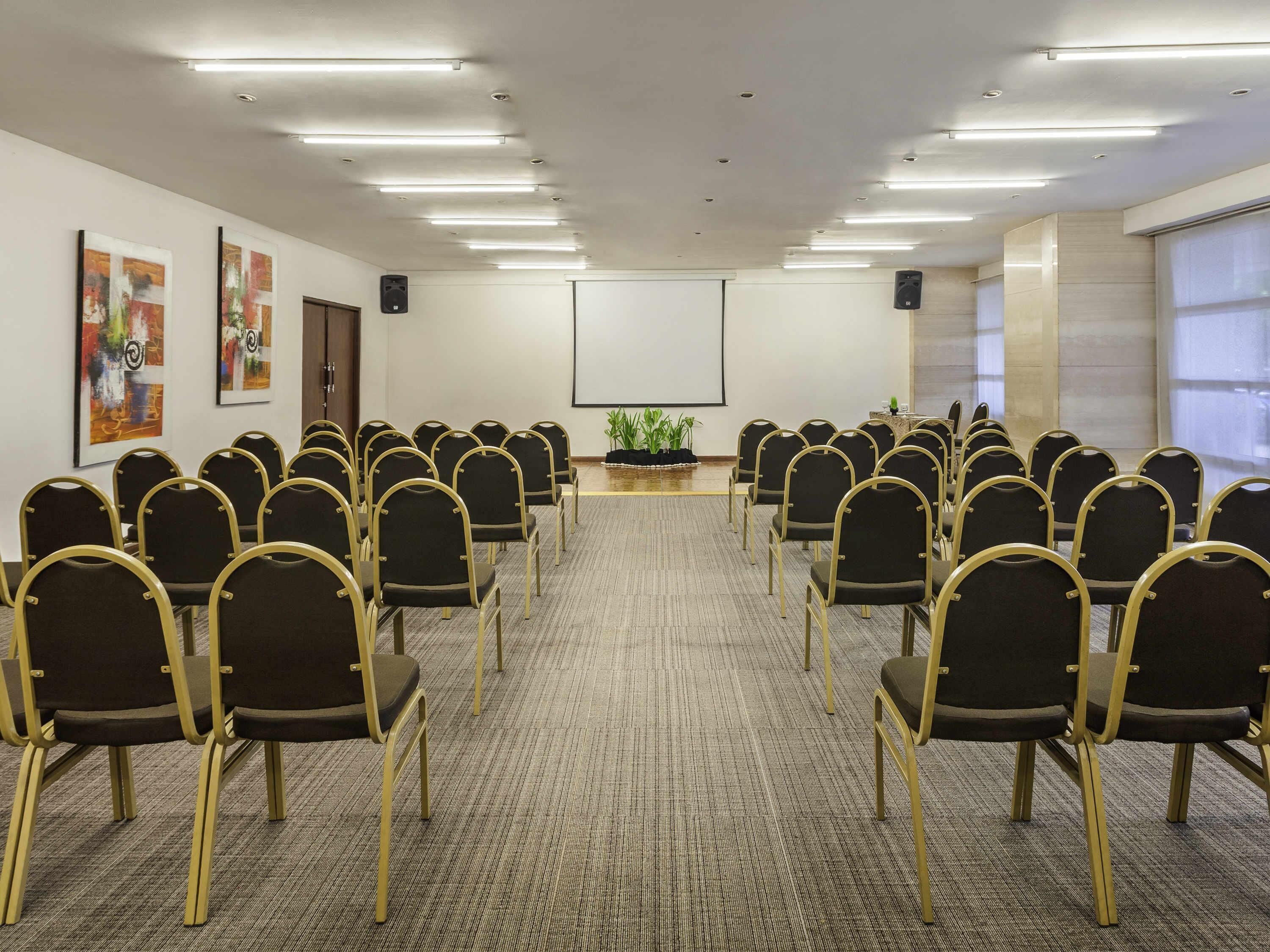 A conference room with rows of chairs, a projector screen, and wall-mounted speakers.