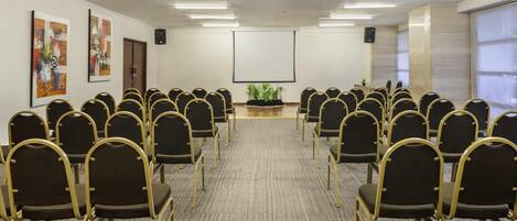 A conference room with rows of chairs, a projector screen, and wall-mounted speakers.