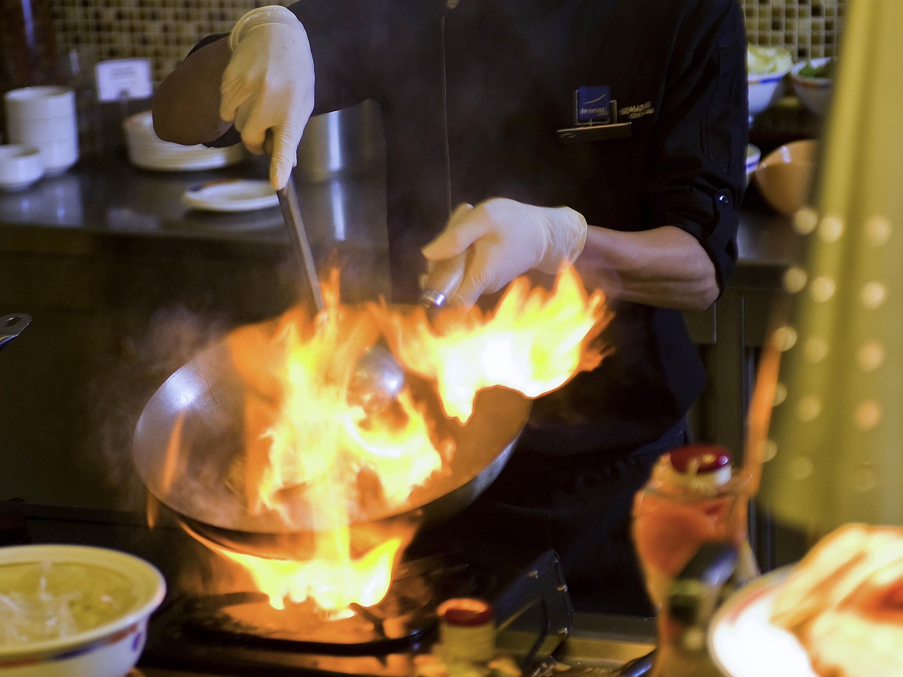 Un cocinero preparando comida en una sartén wok sobre una estufa, rodeado de utensilios y productos frescos.