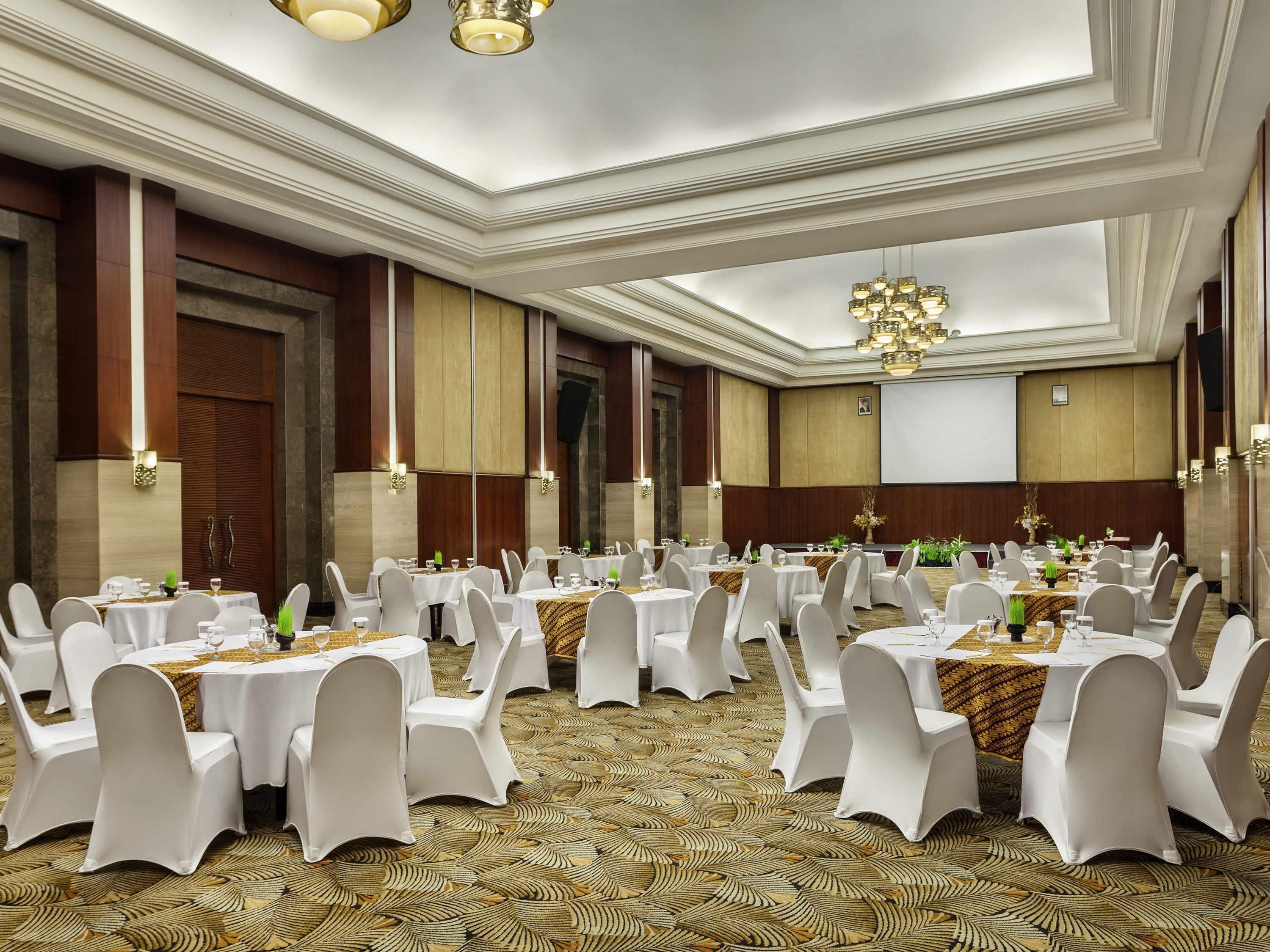 A banquet hall with round tables, white chairs, and a patterned carpet.