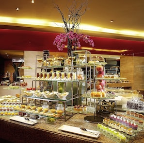 A buffet with various desserts and pastries, a vase with pink flowers, and a red wall in the background.