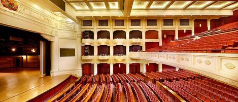 A large auditorium with tiered seating, a stage, and ornate ceiling details.