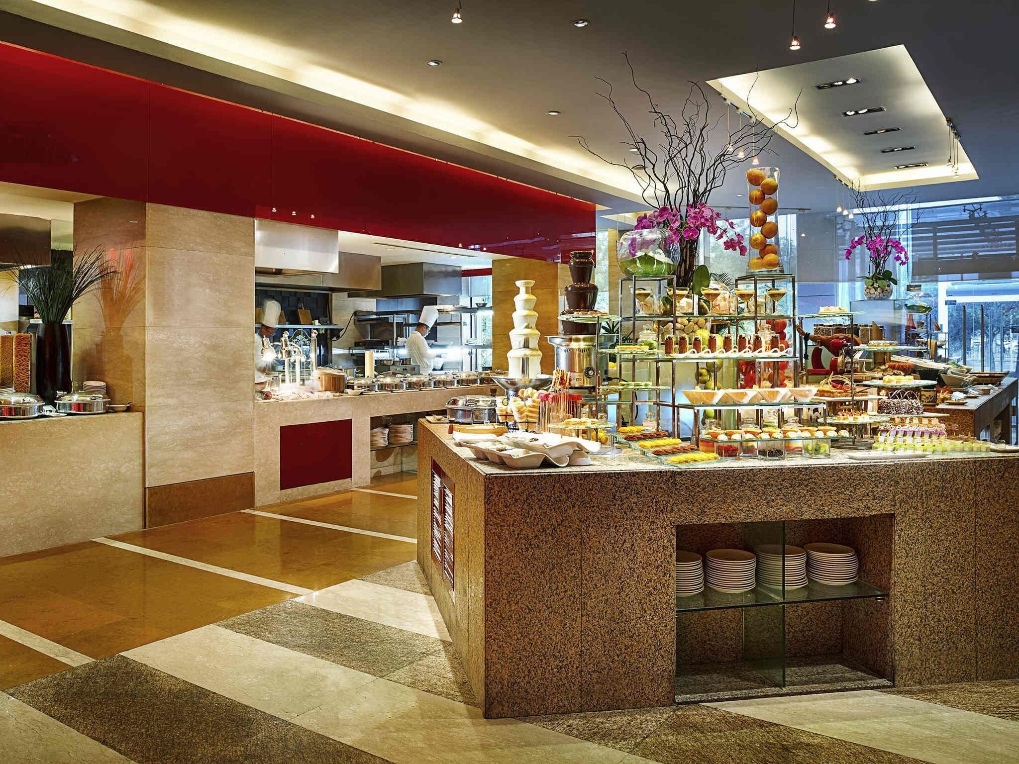 A modern hotel lobby with a central food display, a red accent wall, and a kitchen area in the background.