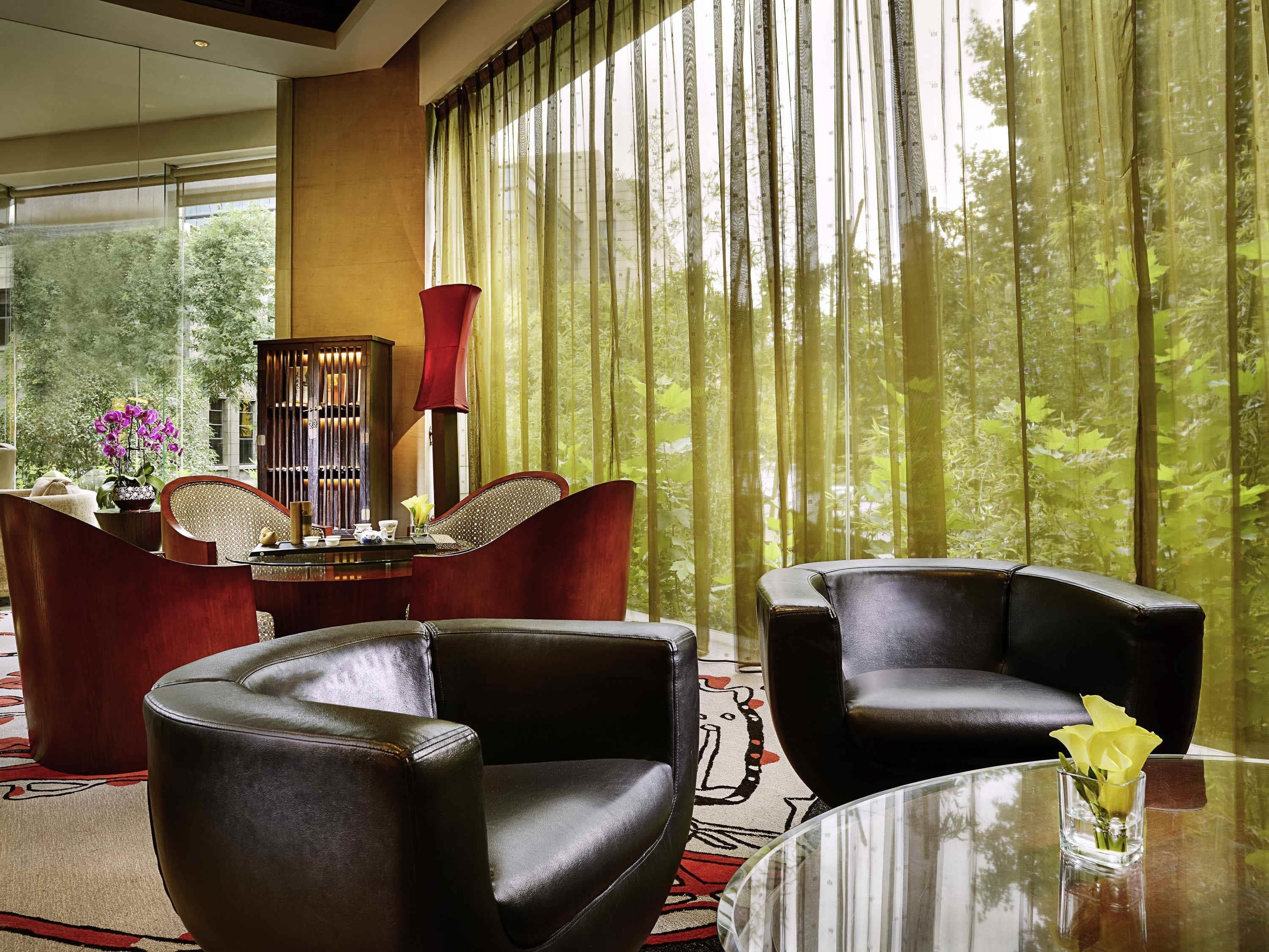A modern hotel room with leather armchairs, a glass coffee table, and a view of a forest through sheer curtains.