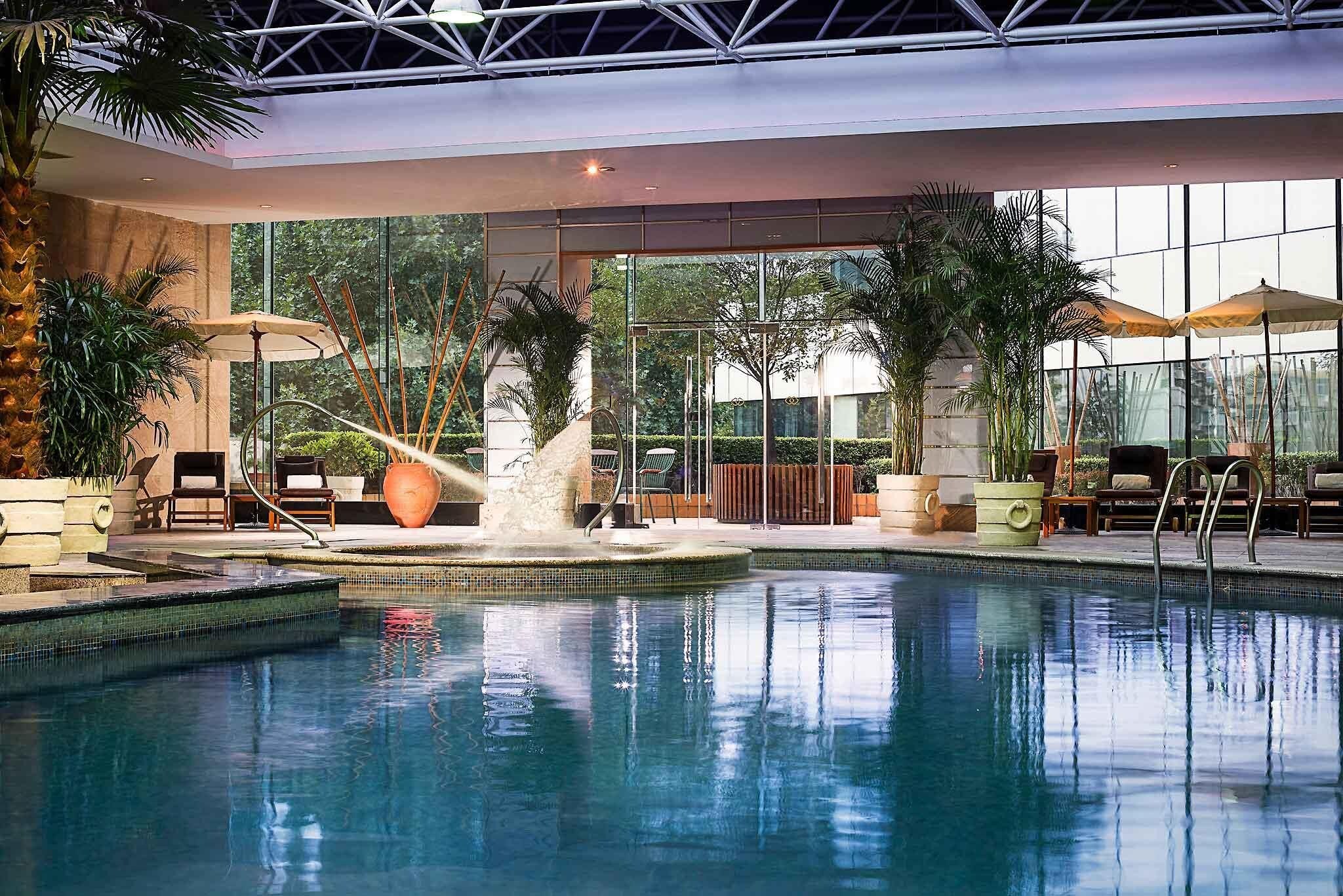 An indoor swimming pool with a water feature, surrounded by lounge chairs and potted plants.