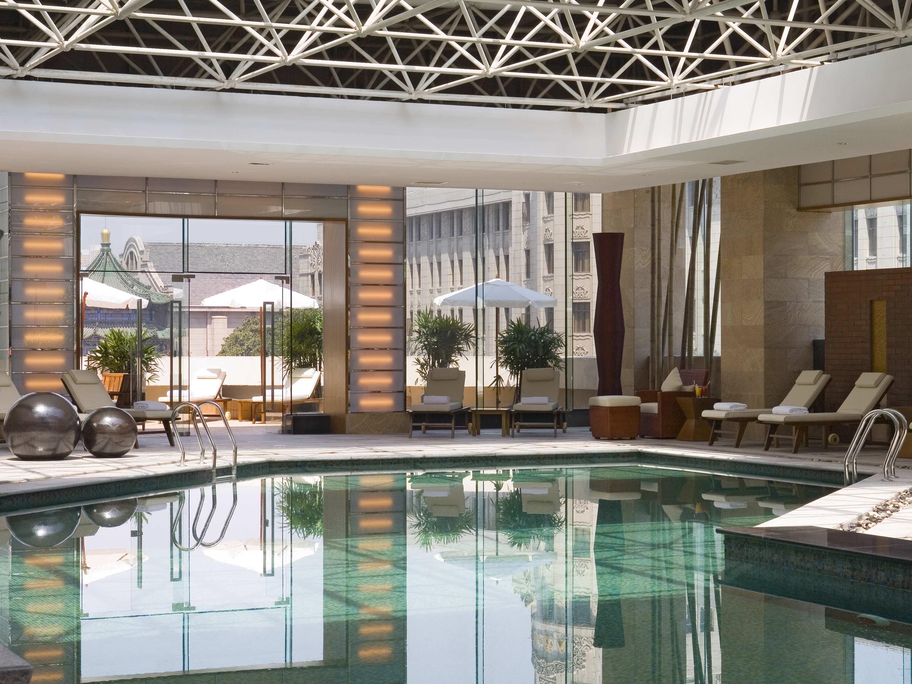A modern indoor pool area with lounge chairs, a glass pool, and a view of traditional architecture outside.