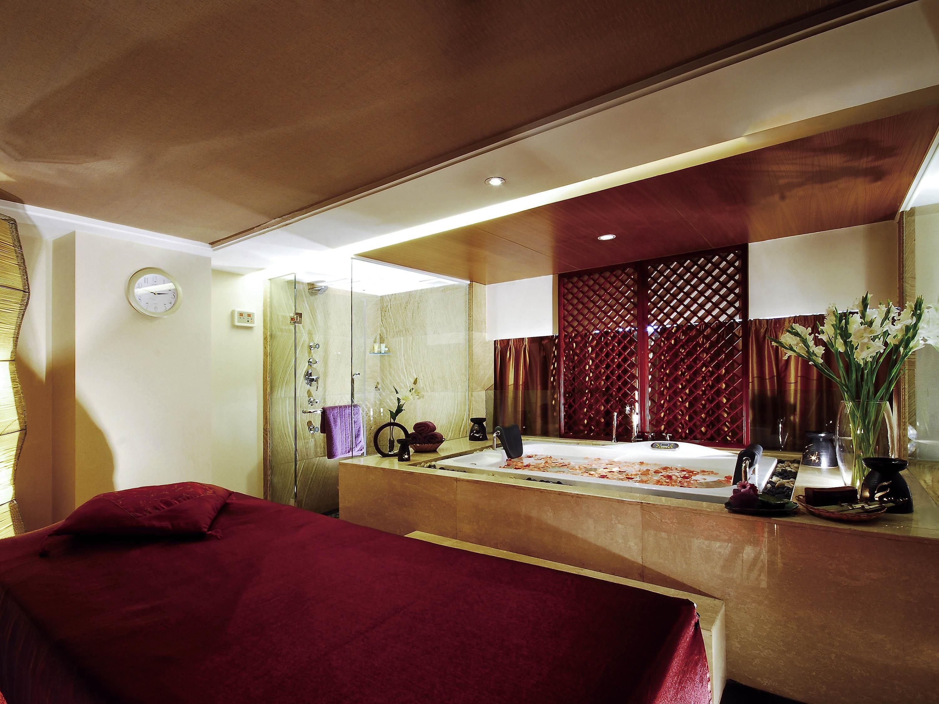 Une chambre d’hôtel avec un grand miroir, un lit recouvert d’une couverture rouge, une baignoire et une coiffeuse avec des articles de toilette.