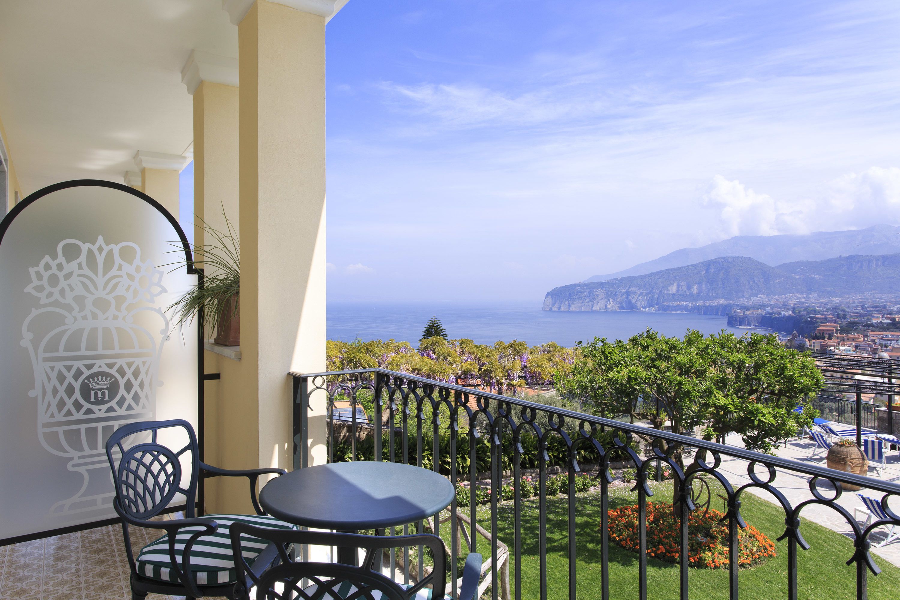 A balcony with a table and chairs, overlooking a scenic view of the sea and mountains.