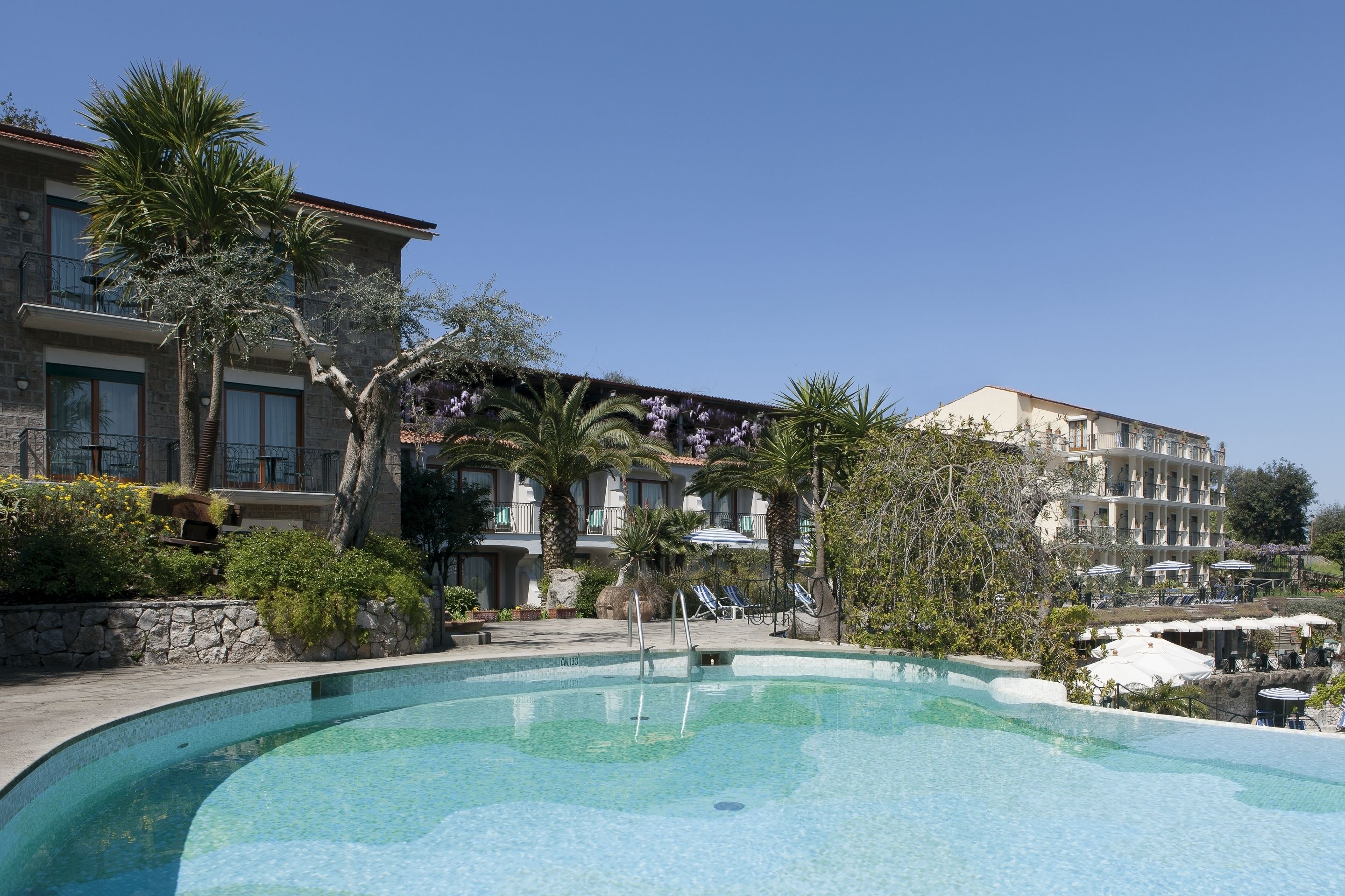 A swimming pool with lounge chairs and umbrellas, surrounded by palm trees and a building with balconies.