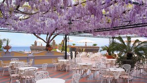 An outdoor patio with white tables and chairs, a view of the sea, and purple flowering trees. - Grand Hotel Capodimonte (Sorrento)